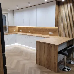 Nowoczesna kuchnia w kształcie litery L z białymi frontami szafek, drewnianym blatem i panelem ściennym, oświetleniem LED pod szafkami wiszącymi, drewnianą ścianką z listew oraz wyspą z dwoma...