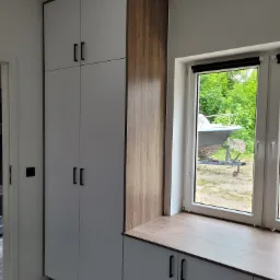 Wysoka szafa z białymi frontami i drewnianymi bokami, zintegrowana z blatem pod oknem, za którym widać przyczepę z łodzią.