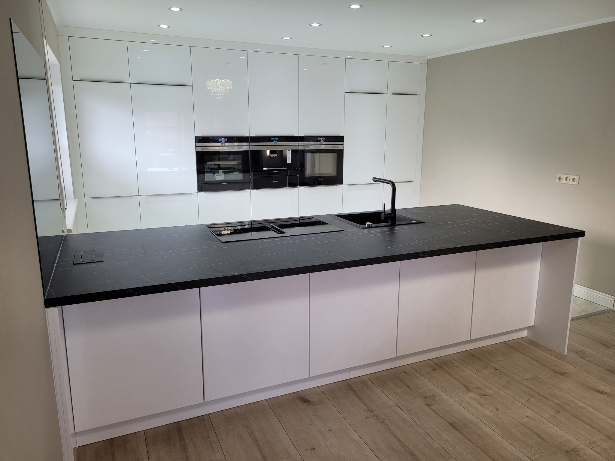 Nowoczesna kuchnia z białymi, lakierowanymi frontami szafek i czarnym blatem z marmurowym wzorem na wyspie kuchennej, oświetlona punktowymi lampami sufitowymi i wiszącym żyrandolem.
