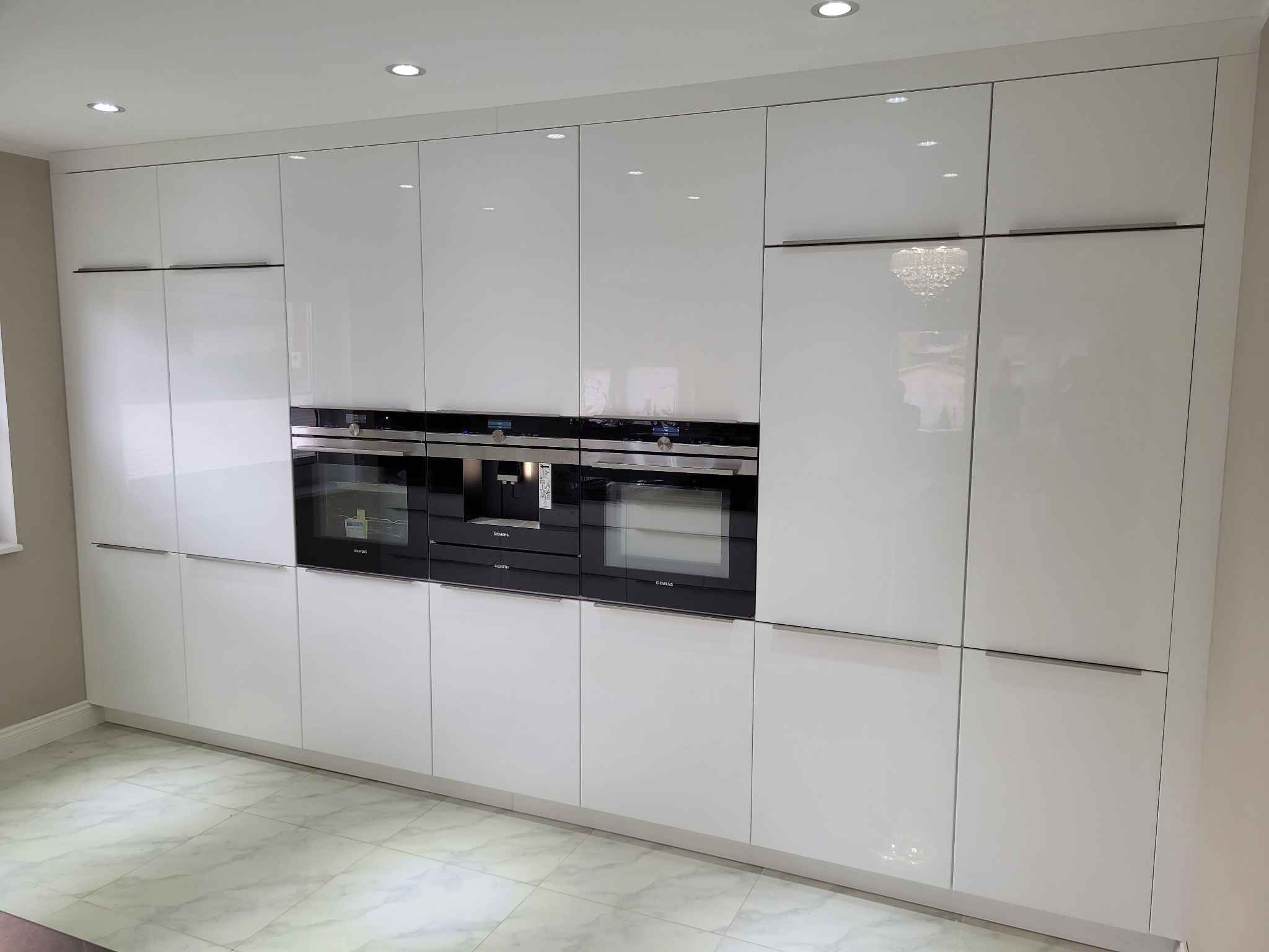 Biała zabudowa kuchenna z połyskiem, z wbudowanymi piekarnikami i ekspresem do kawy marki Siemens, marmurowa podłoga.