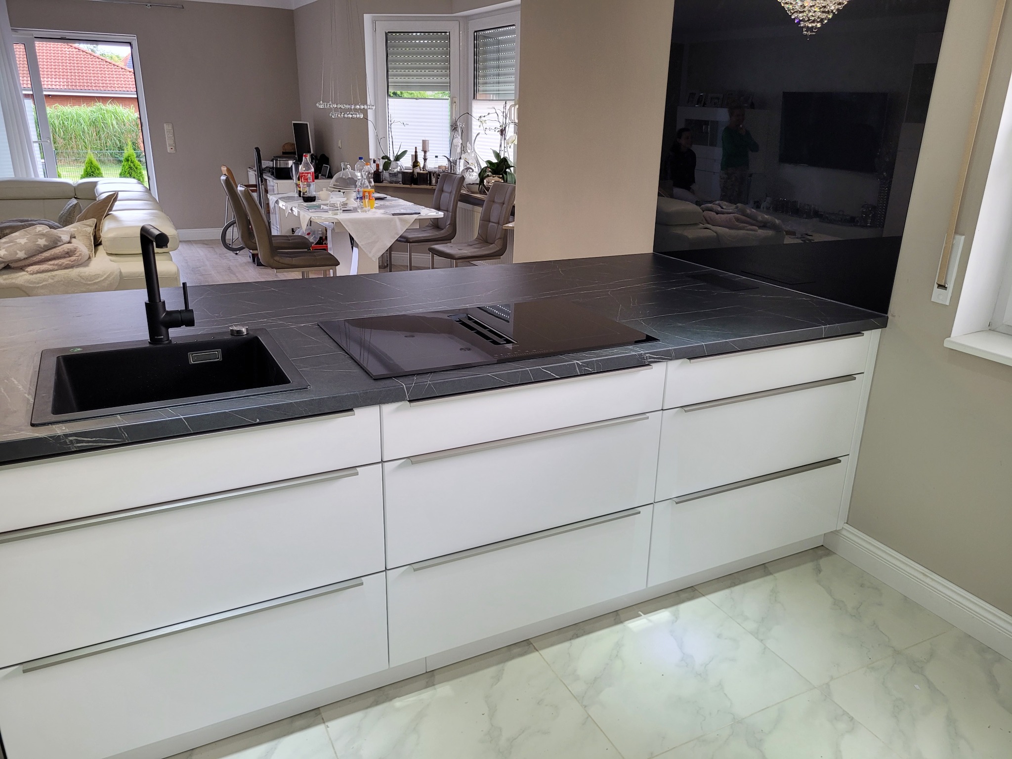 Nowoczesna, biała zabudowa kuchenna z czarnym blatem imitującym marmur i wbudowaną płytą indukcyjną, czarny zlew z baterią, widok na jadalnię i salon w tle.