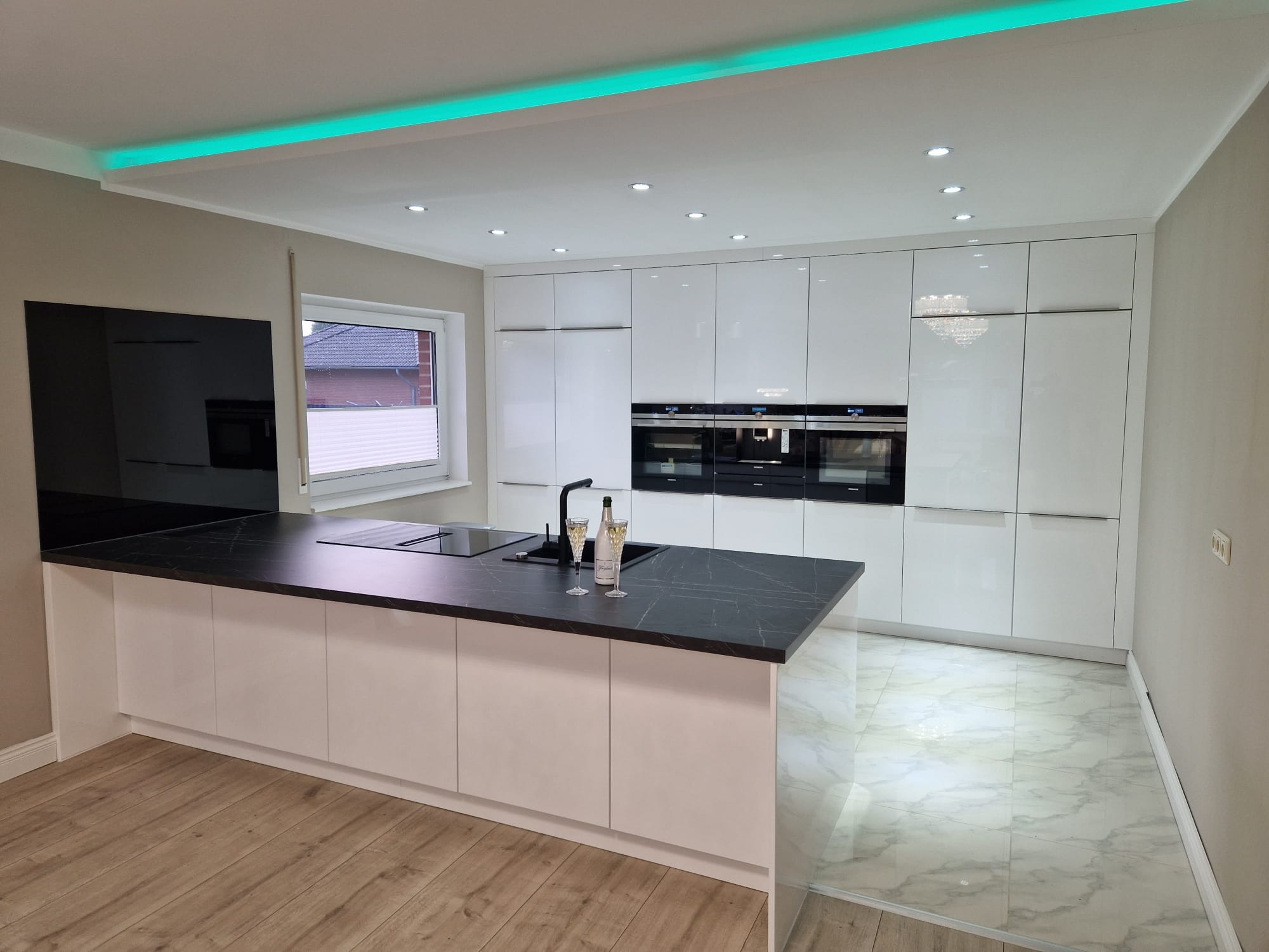 Nowoczesna kuchnia z białymi, lakierowanymi frontami, czarnym blatem i wyspą, oświetleniem LED pod sufitem i szampanem w kieliszkach na blacie.
