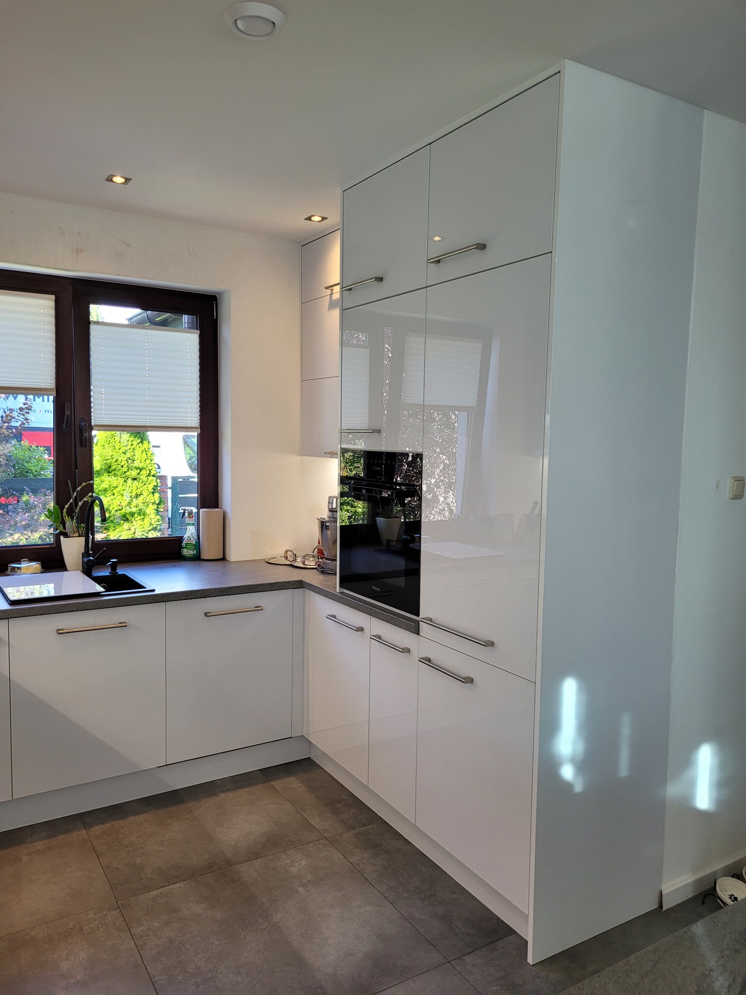 Nowoczesna, biała kuchnia z połyskiem, z oknem wychodzącym na ogród. W tle widoczny wbudowany piekarnik i ciemnoszary blat.