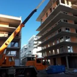 Żuraw samochodowy podnosi materiały budowlane na budowę wielopiętrowego budynku mieszkalnego w słoneczny dzień. Widoczne elementy konstrukcyjne i okna w stanie surowym.