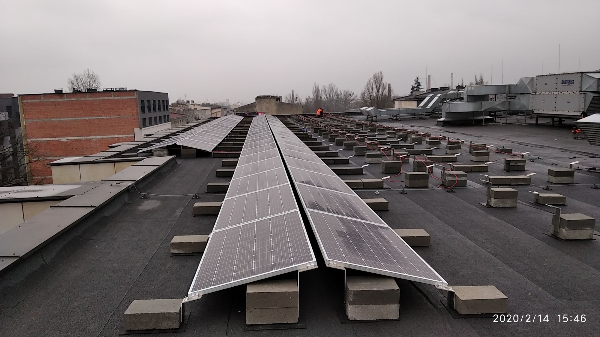 Instalacja fotowoltaiczna na dachu hotelu w Łodzi. Panele zamontowane na betonowych balastach, widoczny pracownik w pomarańczowej kamizelce. W tle budynki przemysłowe i pochmurne niebo.