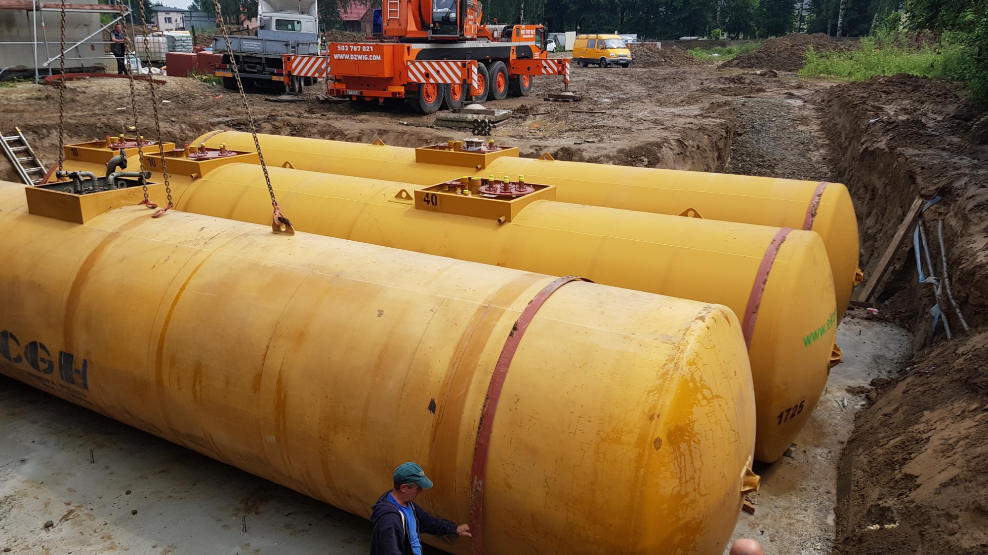 Żółte zbiorniki podziemne układane w wykopie, widoczny żuraw i pracownik w niebieskiej bluzie.