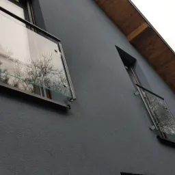 Szklane panele balkonowe z metalowymi mocowaniami na szarej elewacji budynku, odbicie drzew w szkle.