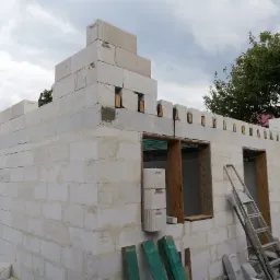 Niedokończony dom z białych bloczków z charakterystycznymi zębami na górze ścian, widoczne drewniane ramy okienne i drabina oparta o ścianę.