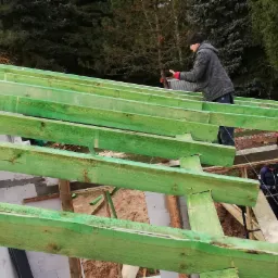 Konstrukcja dachu z zielonych belek, pracownik w czapce i kurtce montuje elementy, widoczny fragment budynku z białych bloczków oraz piła łańcuchowa na dachu.