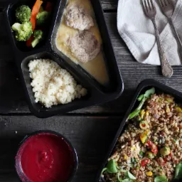 Dwa czarne pojemniki z gotowymi posiłkami: jeden z pulpetami w sosie, kaszą kuskus i warzywami (brokuły, marchew), drugi z kaszą gryczaną z warzywami i ziołami, obok miseczka z sosem buraczkowym...