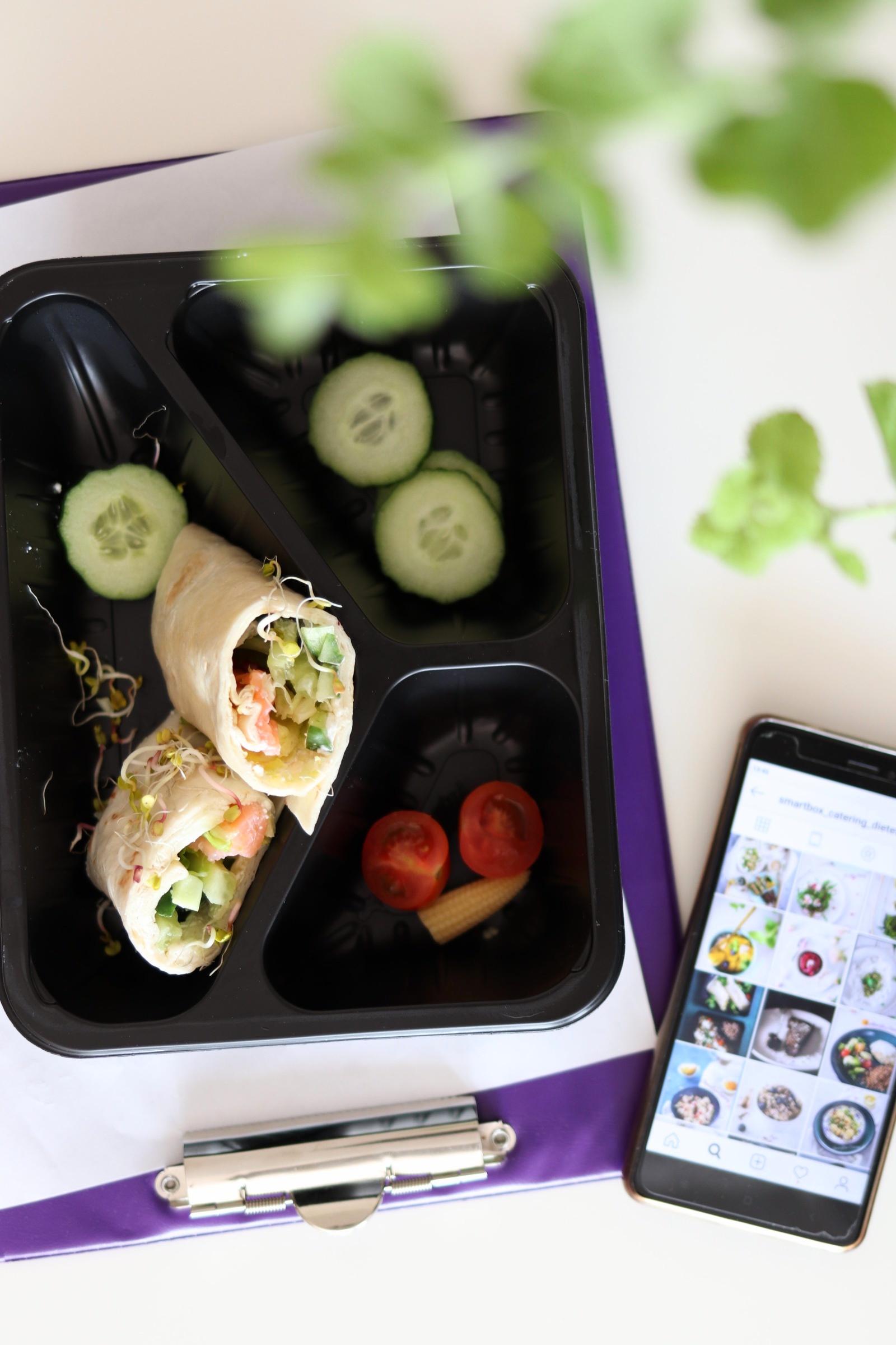 Lunchbox z wrapami, ogórkiem i pomidorkami koktajlowymi, obok telefon z widocznym profilem cateringowym na Instagramie, ujęcie z góry.