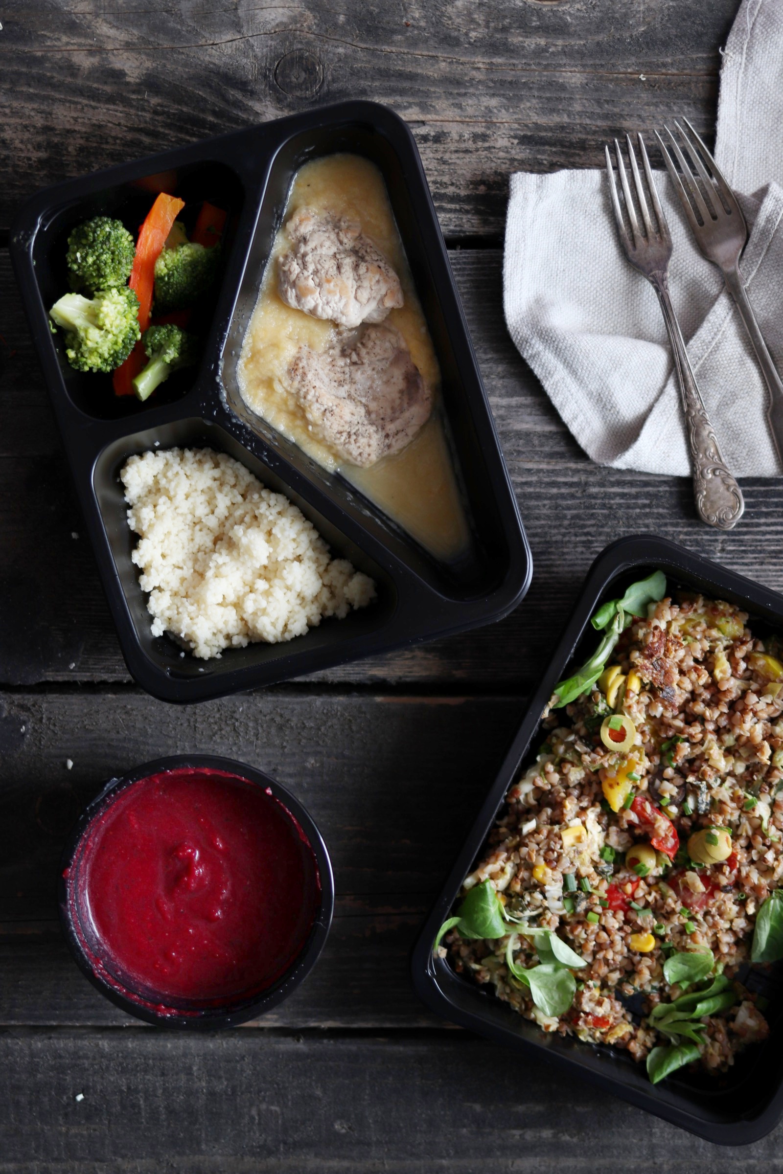 Dwa czarne pojemniki z gotowymi posiłkami: jeden z pulpetami w sosie, kaszą kuskus i warzywami (brokuły, marchew), drugi z kaszą gryczaną z warzywami i ziołami, obok miseczka z sosem buraczkowym...