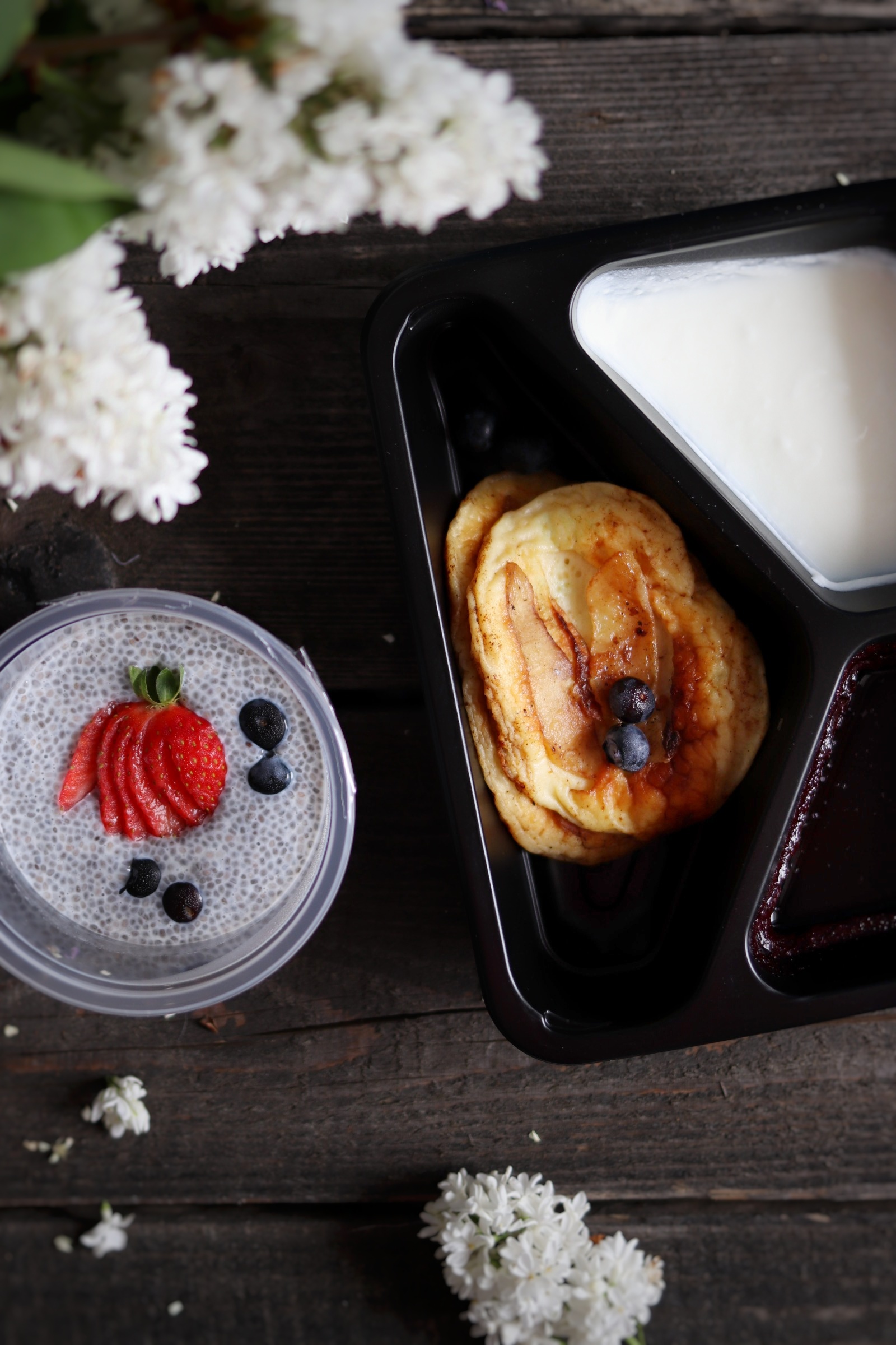 Pudełko z naleśnikami z jabłkami i jagodami, jogurtem i dżemem obok chia puddingu z truskawkami i jagodami, na tle drewnianego stołu z gałązkami bzu.