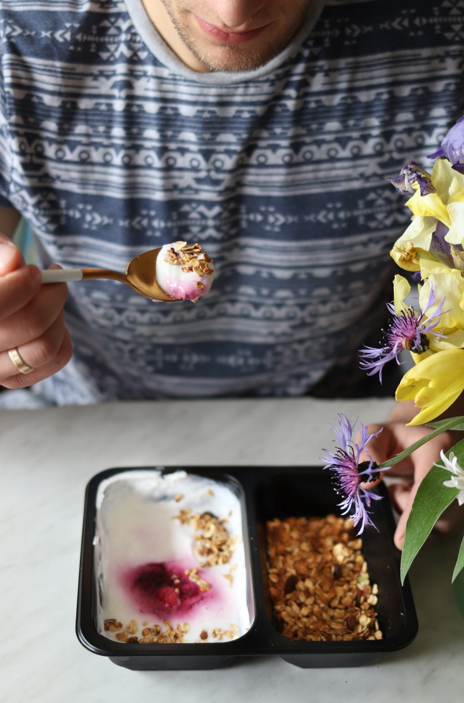 Mężczyzna z łyżką jogurtu z granolą i owocami, obok pudełko z dwoma przegródkami, w jednej jogurt z owocami i granolą, w drugiej sama granola, dekoracja z kwiatów w tle.
