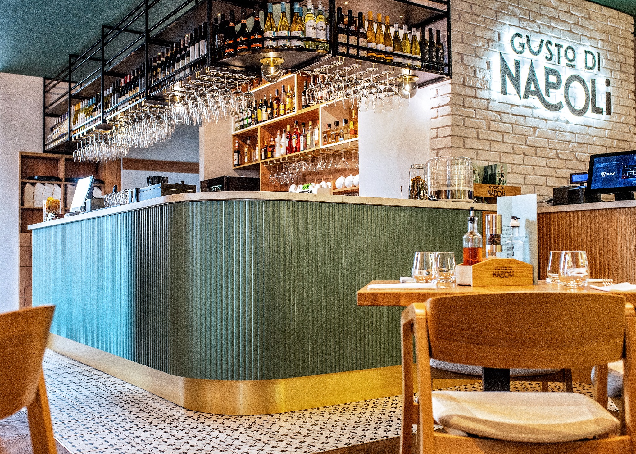 Wnętrze baru restauracji z zielonym, żłobkowanym barem z mosiężnym wykończeniem, półkami z alkoholem i szklankami, oraz stolikiem z nakryciem i napisem neonowym 'Gusto di Napoli' na ścianie z cegły.