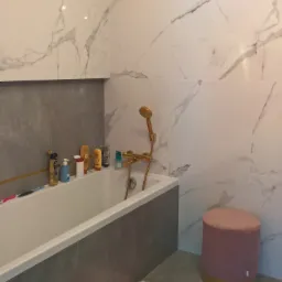 Nowoczesna łazienka z marmurowymi płytkami, złotą armaturą i pudrowo-różowymi akcentami: puf i dywanik.