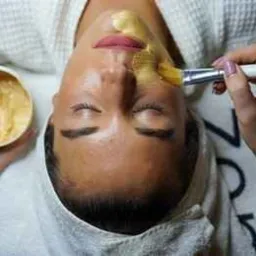 Nakładanie złotej maski pędzlem na twarz klientki w salonie w Pile. Klientka leży z zamkniętymi oczami, na głowie ma biały ręcznik.