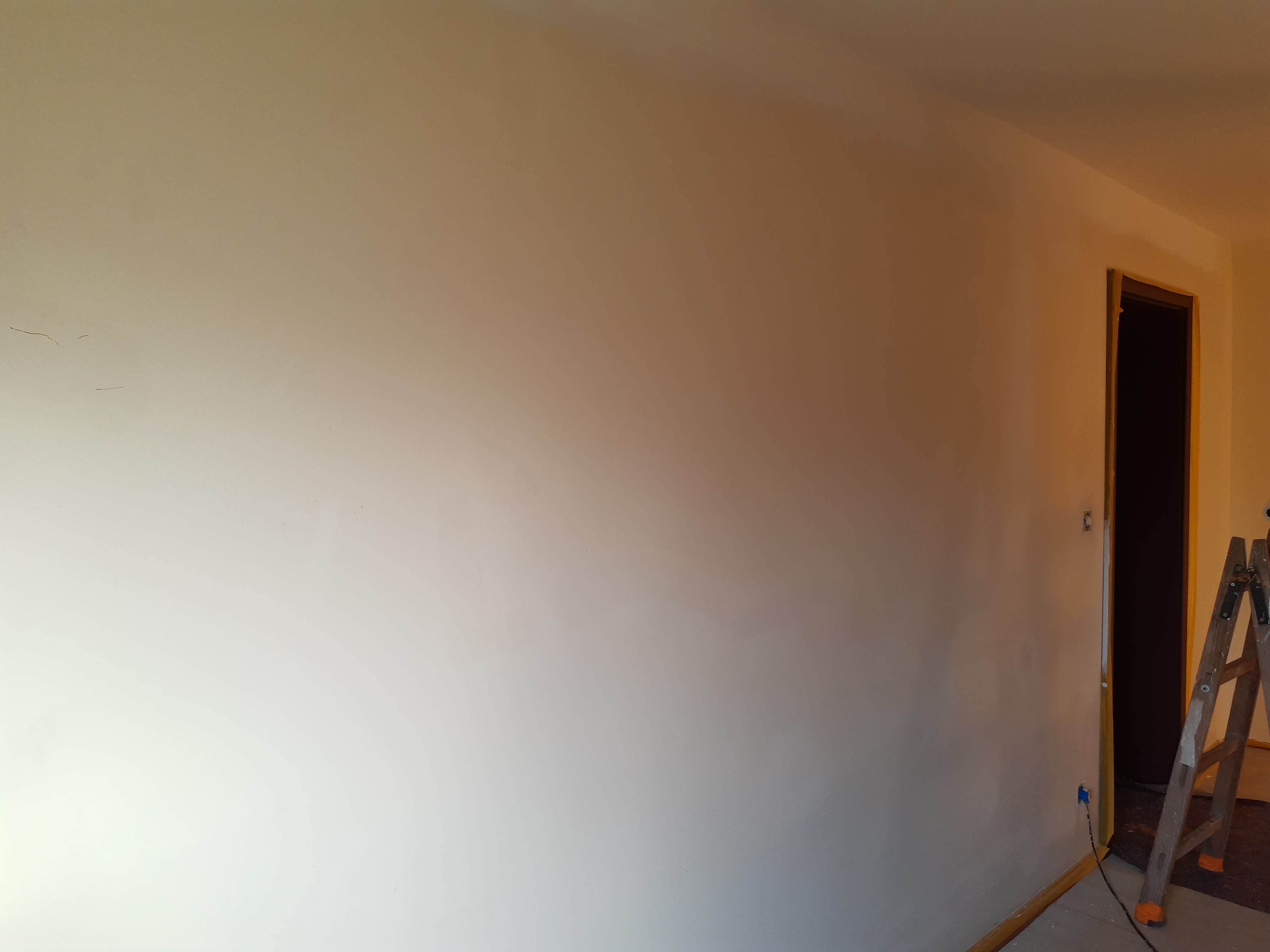 Świeżo pomalowana ściana w odcieniach beżu, widoczne drobne niedoskonałości i ślady po wcześniejszych pracach, w tle otwór drzwiowy i drabina malarska.