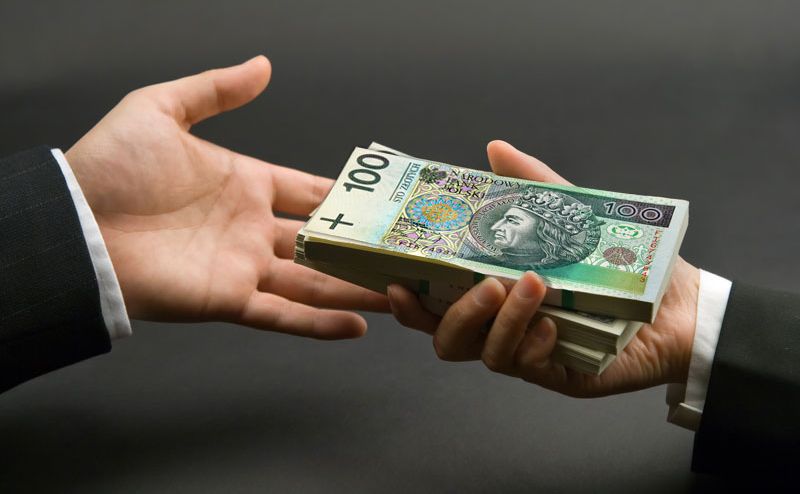 Dwie dłonie wymieniające się plikiem banknotów 100 zł na ciemnym tle, widoczny fragment marynarki.