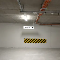 Oznakowanie wyjazdu z parkingu podziemnego: tablica z napisem 'Wyjazd' i strzałką, żółto-czarne pasy ostrzegawcze na ścianie, widoczne instalacje pod sufitem.