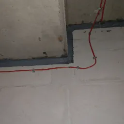 Uszczelnienie szarej fugi między betonowymi elementami ściany i sufitu, z poprowadzonymi czerwonymi przewodami elektrycznymi mocowanymi metalowymi uchwytami.