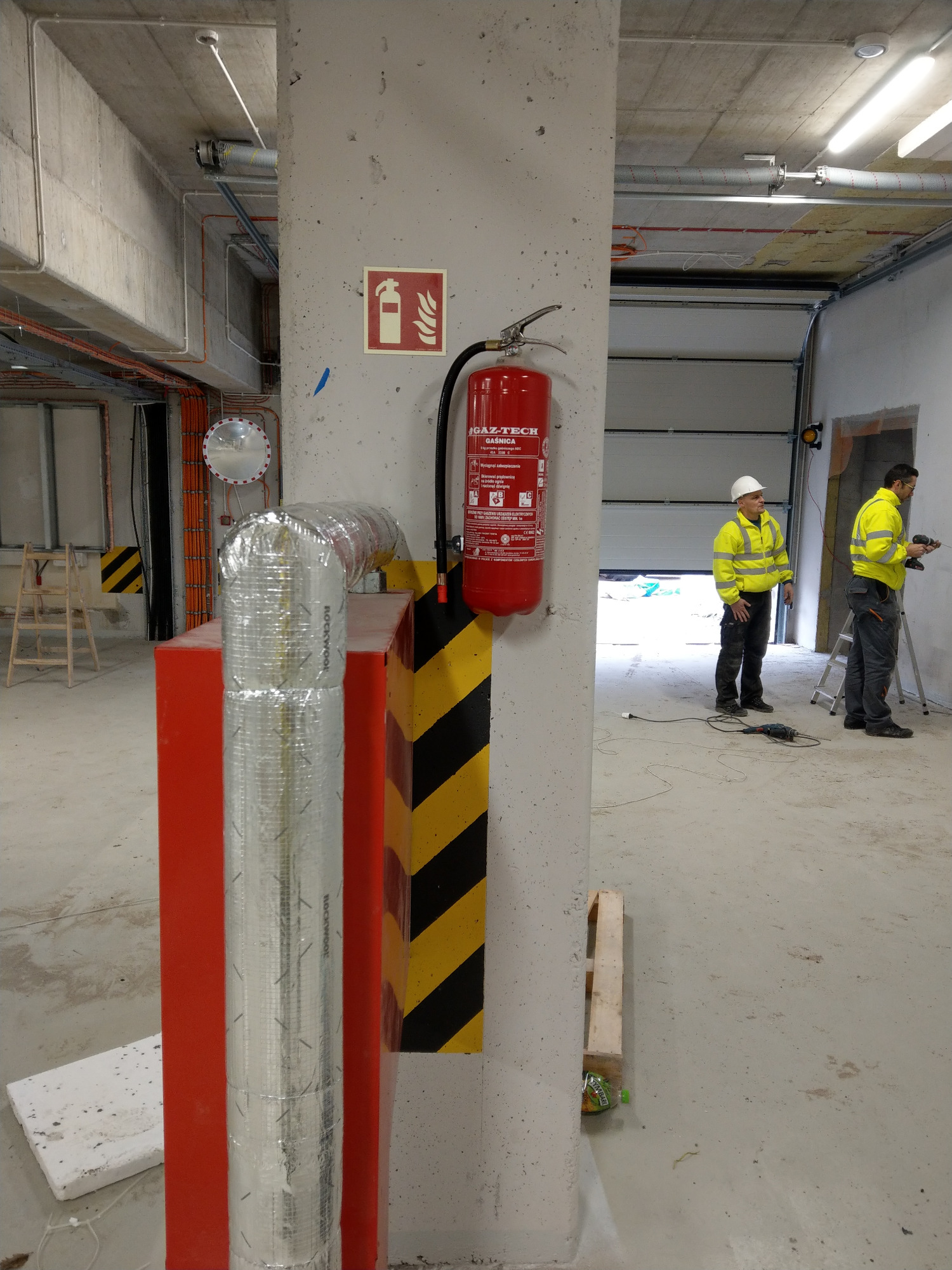 Instalacja systemu przeciwpożarowego w hali: gaśnica na betonowym słupie z oznaczeniami ostrzegawczymi, w tle pracownicy w odblaskowych kurtkach montujący instalację.