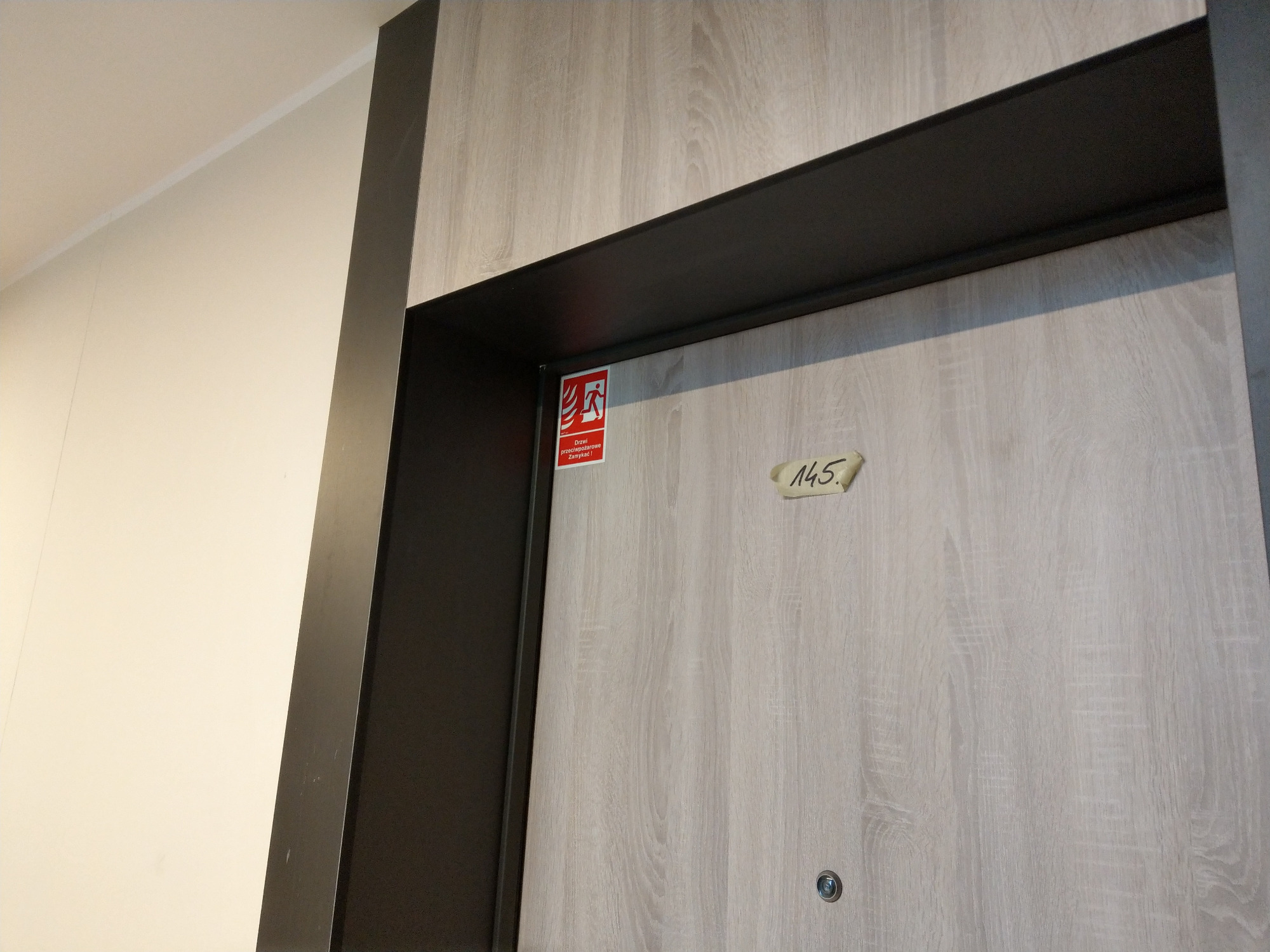 Drewnopodobne drzwi z czarną ościeżnicą, naklejką z numerem 145 i znakiem ewakuacyjnym w lewym górnym rogu.