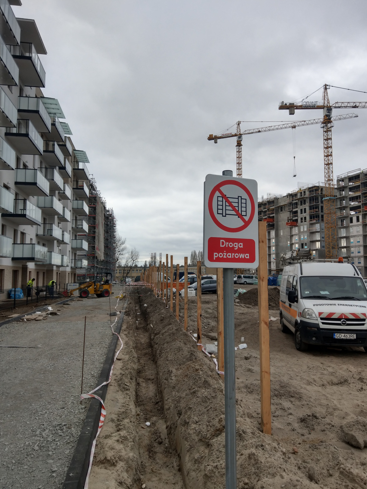 Plac budowy z nowym budynkiem wielorodzinnym w tle, znak 'Droga pożarowa' na pierwszym planie, wykop w ziemi i pracujący robotnicy.