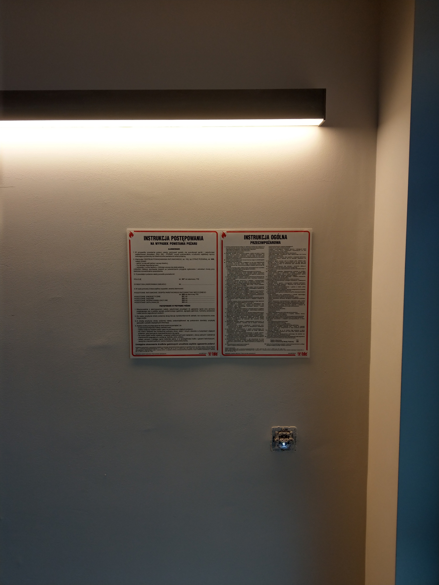 Dwie instrukcje przeciwpożarowe, jedna dotycząca postępowania w razie pożaru, druga ogólna, umieszczone obok siebie na ścianie wewnątrz budynku, pod oświetleniem.
