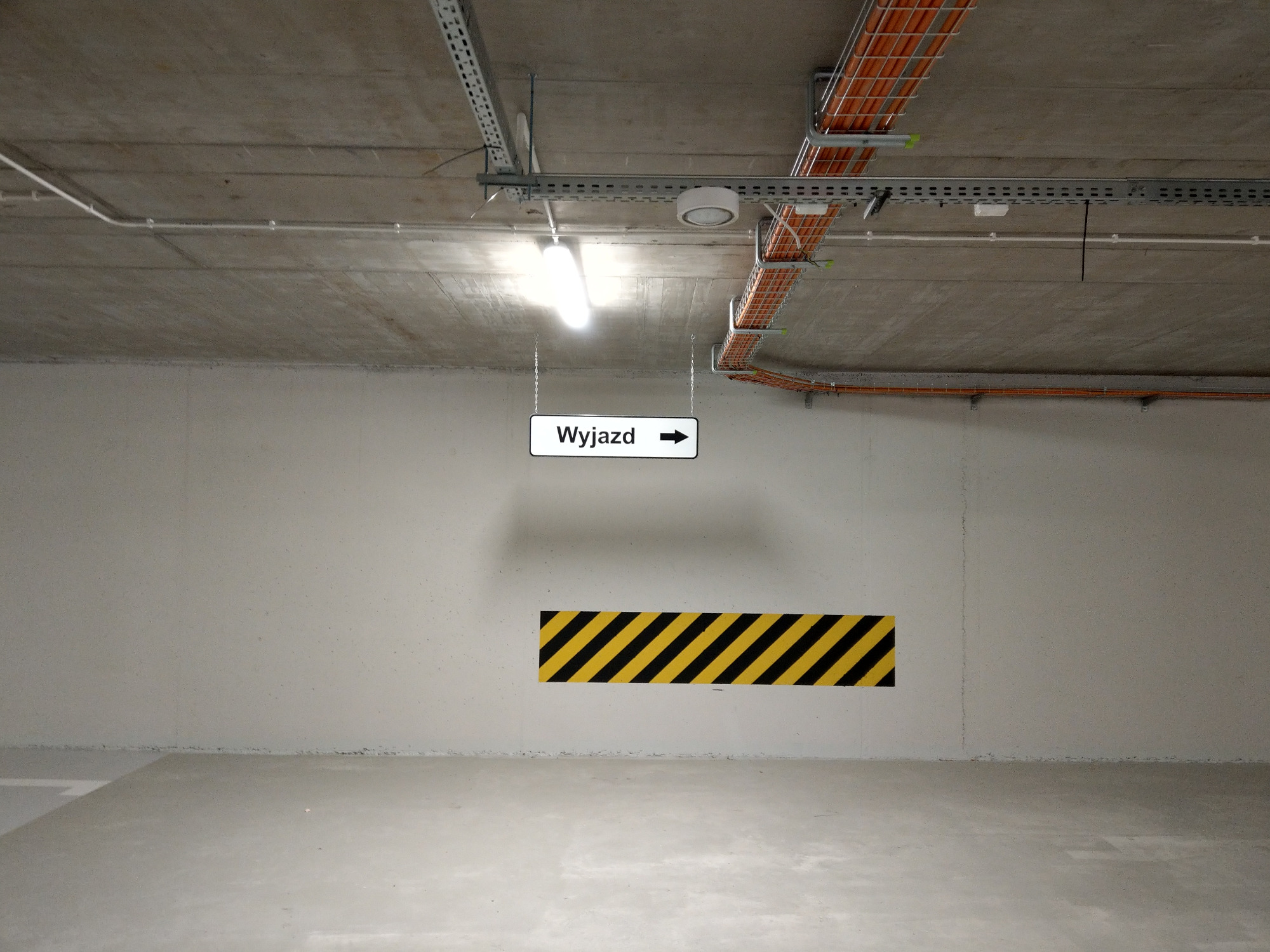 Oznakowanie wyjazdu z parkingu podziemnego: tablica z napisem 'Wyjazd' i strzałką, żółto-czarne pasy ostrzegawcze na ścianie, widoczne instalacje pod sufitem.