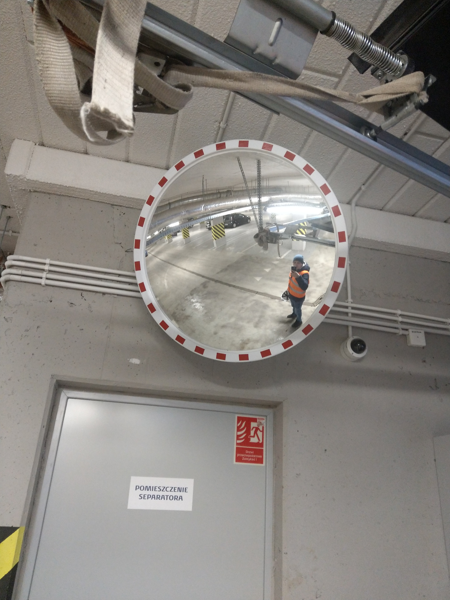 Okrągłe lustro drogowe z czerwoną obwódką, zamontowane w garażu podziemnym, odbijające sylwetkę osoby w kamizelce odblaskowej i niebieskiej czapce.