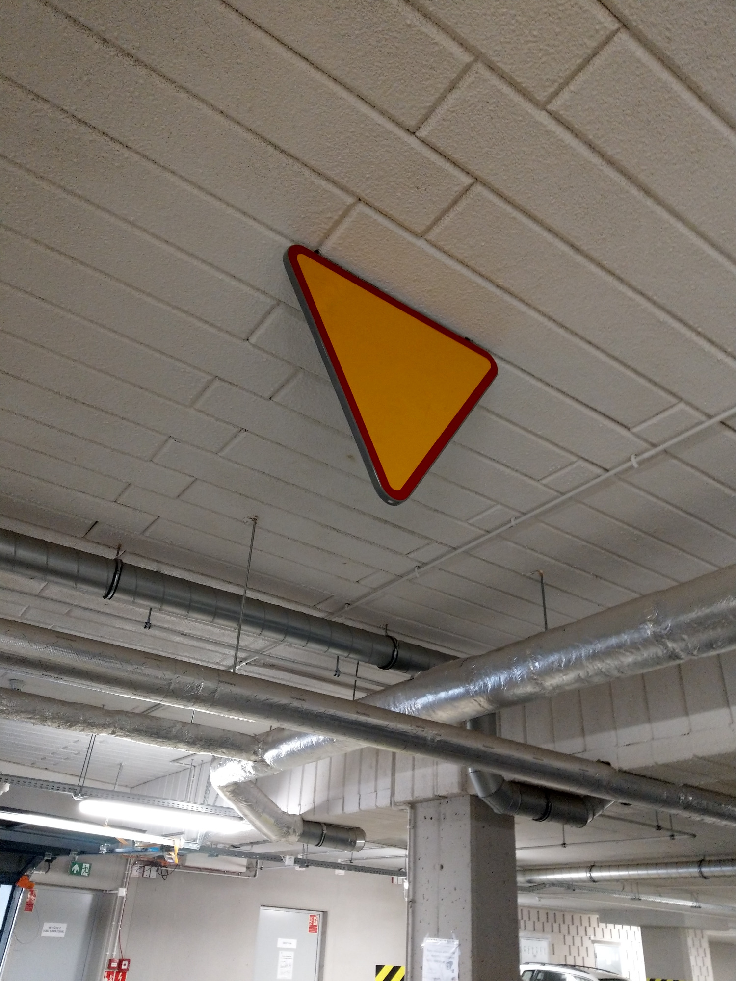 Żółty znak ostrzegawczy w czerwonej obwódce, przymocowany do sufitu parkingu podziemnego, widoczne rury wentylacyjne.