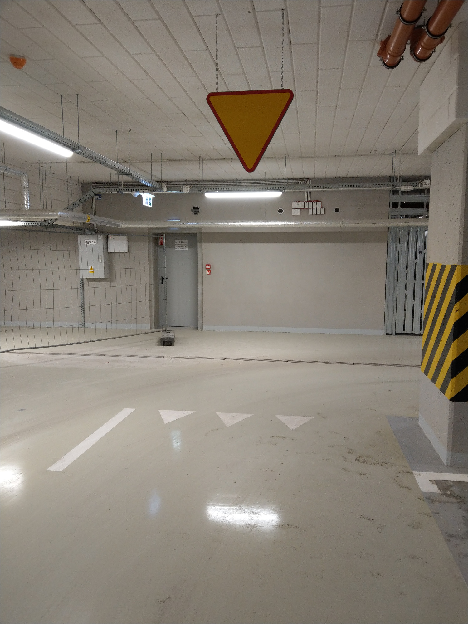 Oznakowanie poziome na parkingu podziemnym z wiszącym znakiem ostrzegawczym, widoczne instalacje pod sufitem i zabezpieczenie narożnika kolumny w czarno-żółte pasy.