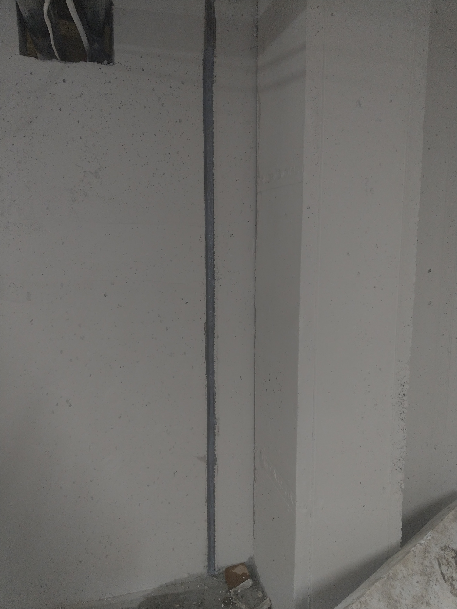 Szara ściana z betonu komórkowego z pionowym rowkiem wypełnionym szarą masą uszczelniającą i widocznym otworem instalacyjnym z wystającymi kablami.