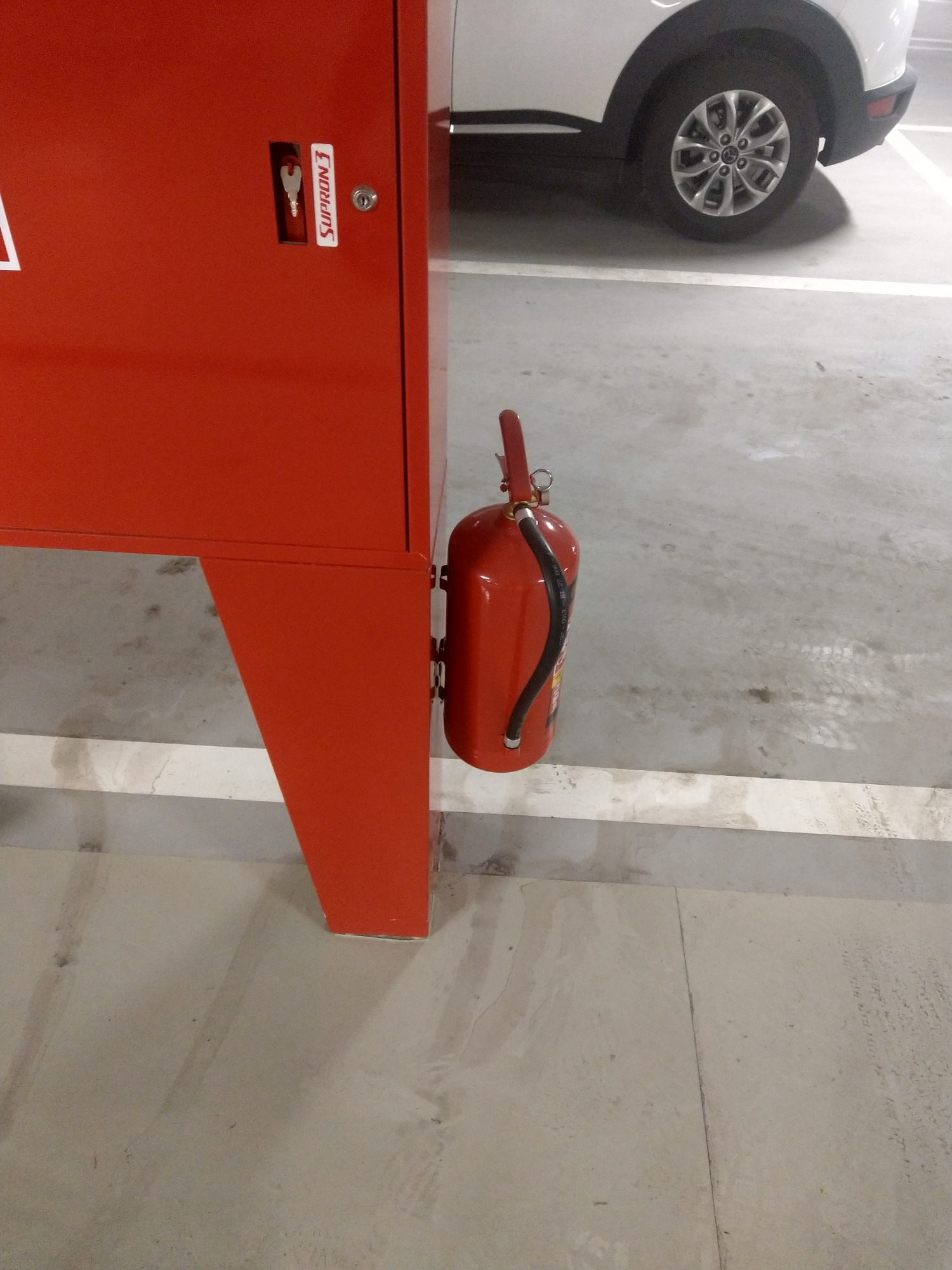 Czerwona szafka z kluczem i gaśnica przymocowana do boku, na tle betonowej podłogi parkingu i fragmentu białego samochodu.