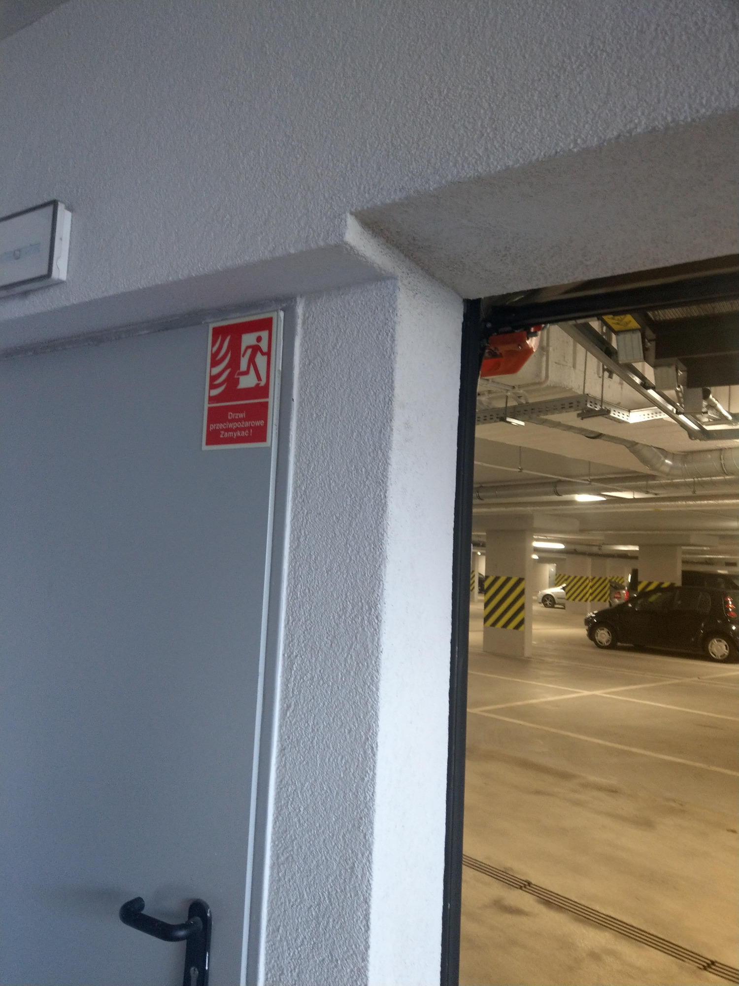 Otwarcie drzwi przeciwpożarowych w garażu podziemnym, widoczny znak ewakuacyjny na szarych drzwiach, oświetlony garaż z samochodami w tle.