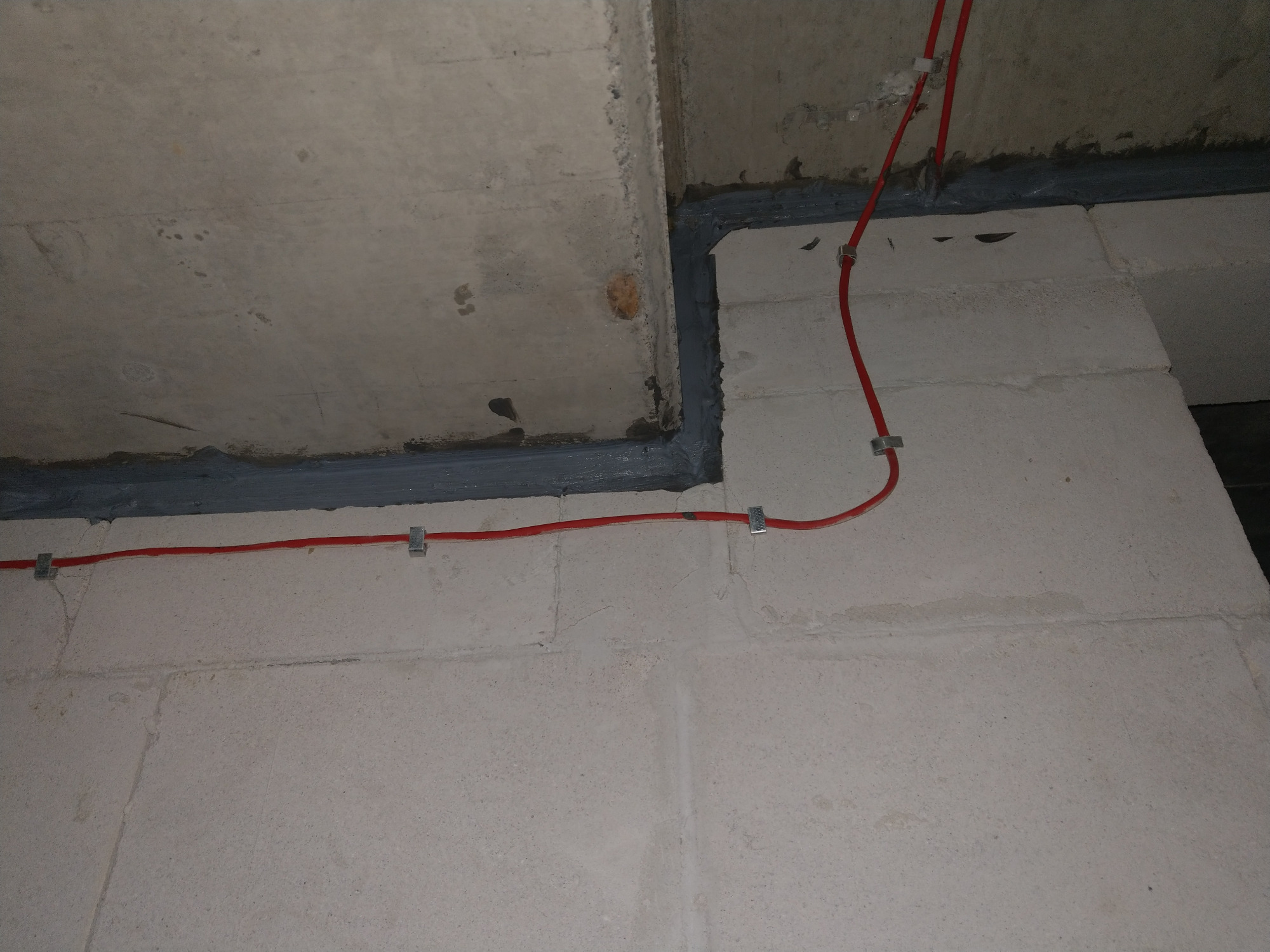 Uszczelnienie szarej fugi między betonowymi elementami ściany i sufitu, z poprowadzonymi czerwonymi przewodami elektrycznymi mocowanymi metalowymi uchwytami.