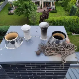 Widok z góry na cztery kominy na dachu, z akcesoriami do czyszczenia: szczotka druciana, obciążnik i lina, na tle zadbanego ogrodu.