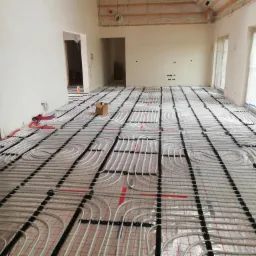 Rozległy pokój w trakcie instalacji ogrzewania podłogowego, widoczne białe rury ułożone na folii z czerwoną siatką, czarne elementy mocujące, surowy stan ścian i sufitu, karton na podłodze.