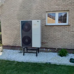 Biała pompa ciepła Vaillant zamontowana na czarnym stelażu przy ścianie budynku z cegły, obok okna z odbiciem nieba i drzew, otoczona białym żwirem i trawnikiem.