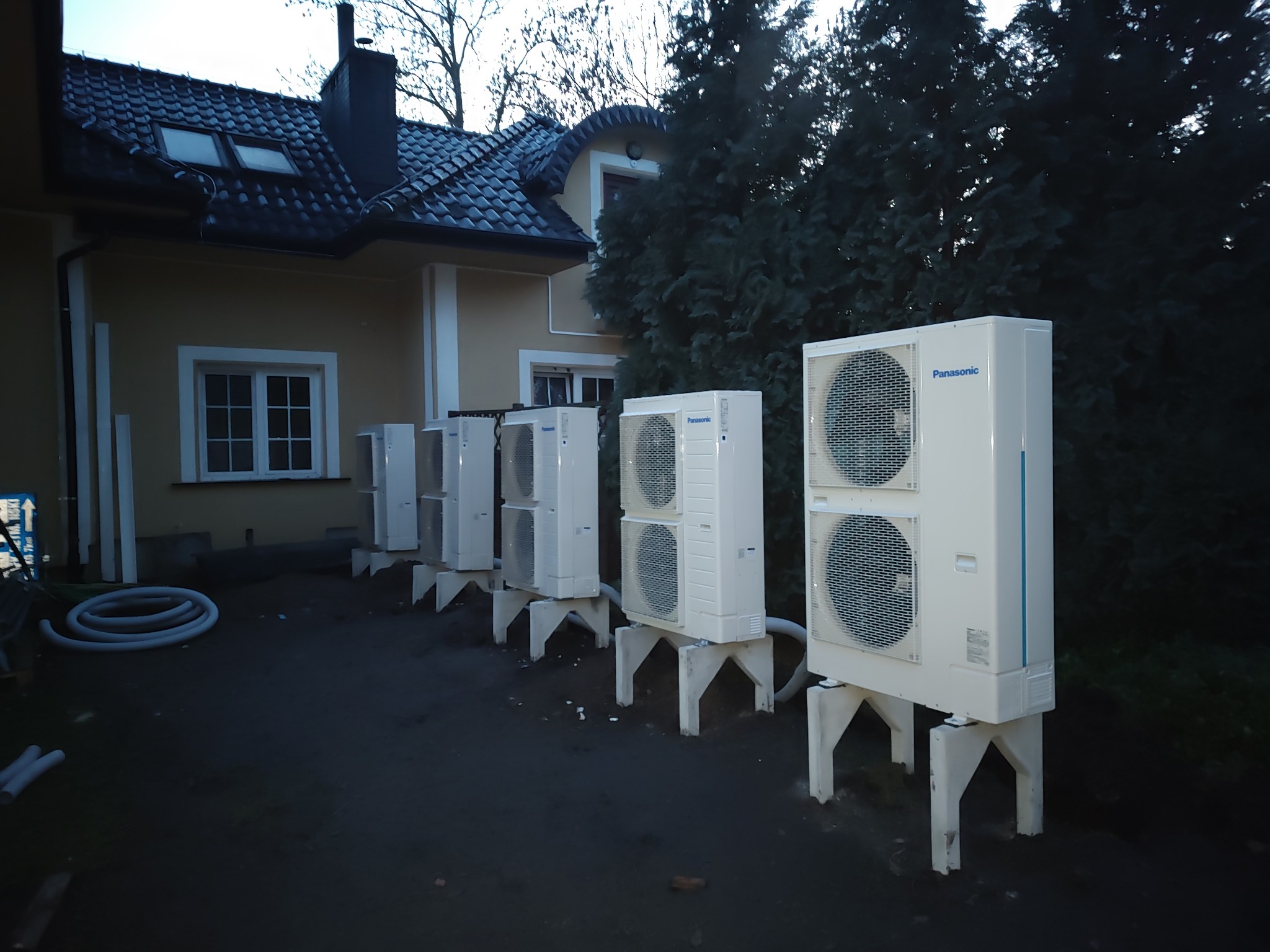 Szereg białych, zewnętrznych jednostek pomp ciepła marki Panasonic ustawionych na betonowych podstawach przed budynkiem mieszkalnym z ciemną dachówką i iglastymi drzewami w tle.