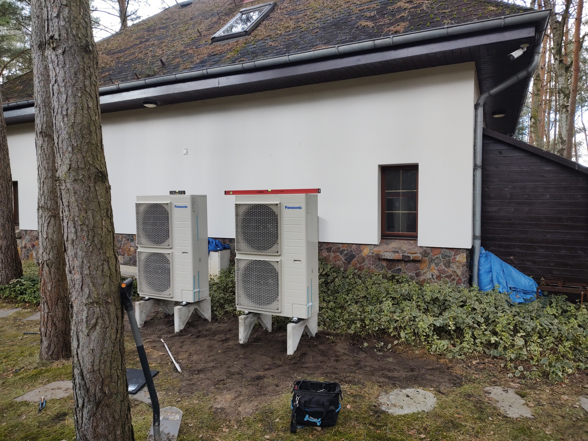 Dwie jednostki zewnętrzne pompy ciepła Panasonic ustawione na betonowych podstawach przed białym budynkiem z dachem porośniętym mchem, widoczne narzędzia i torba monterska.