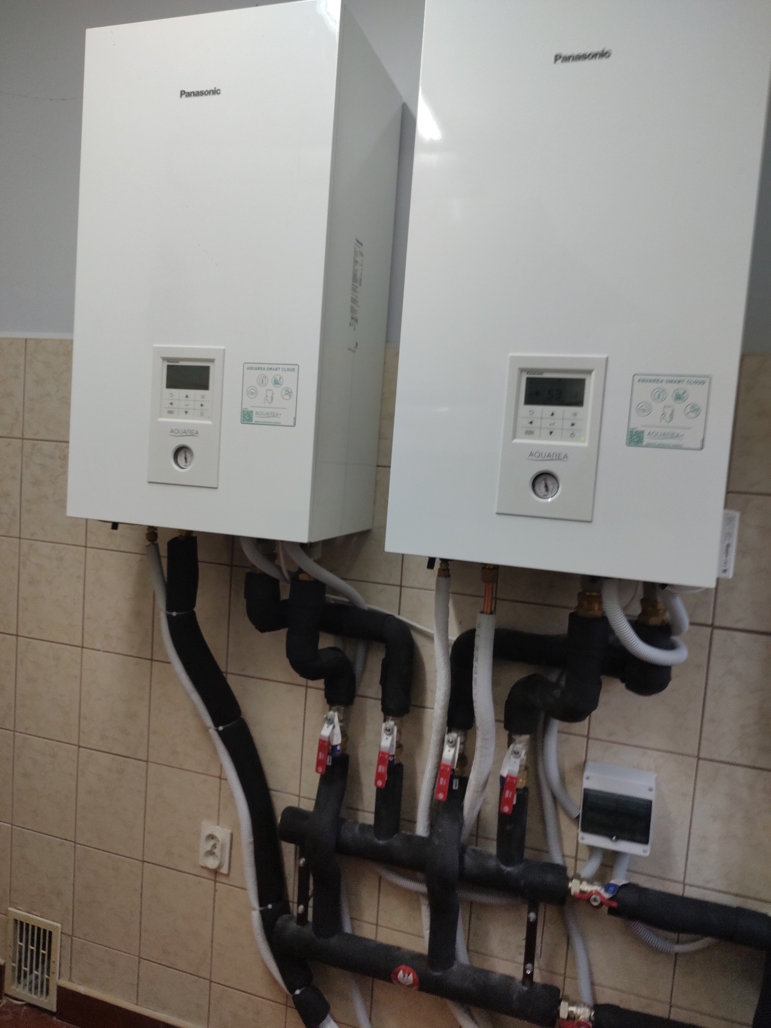 Dwa białe, wiszące moduły Panasonic Aquarea Smart Cloud z widocznymi wyświetlaczami i rurami, zainstalowane na ścianie z beżowych płytek.