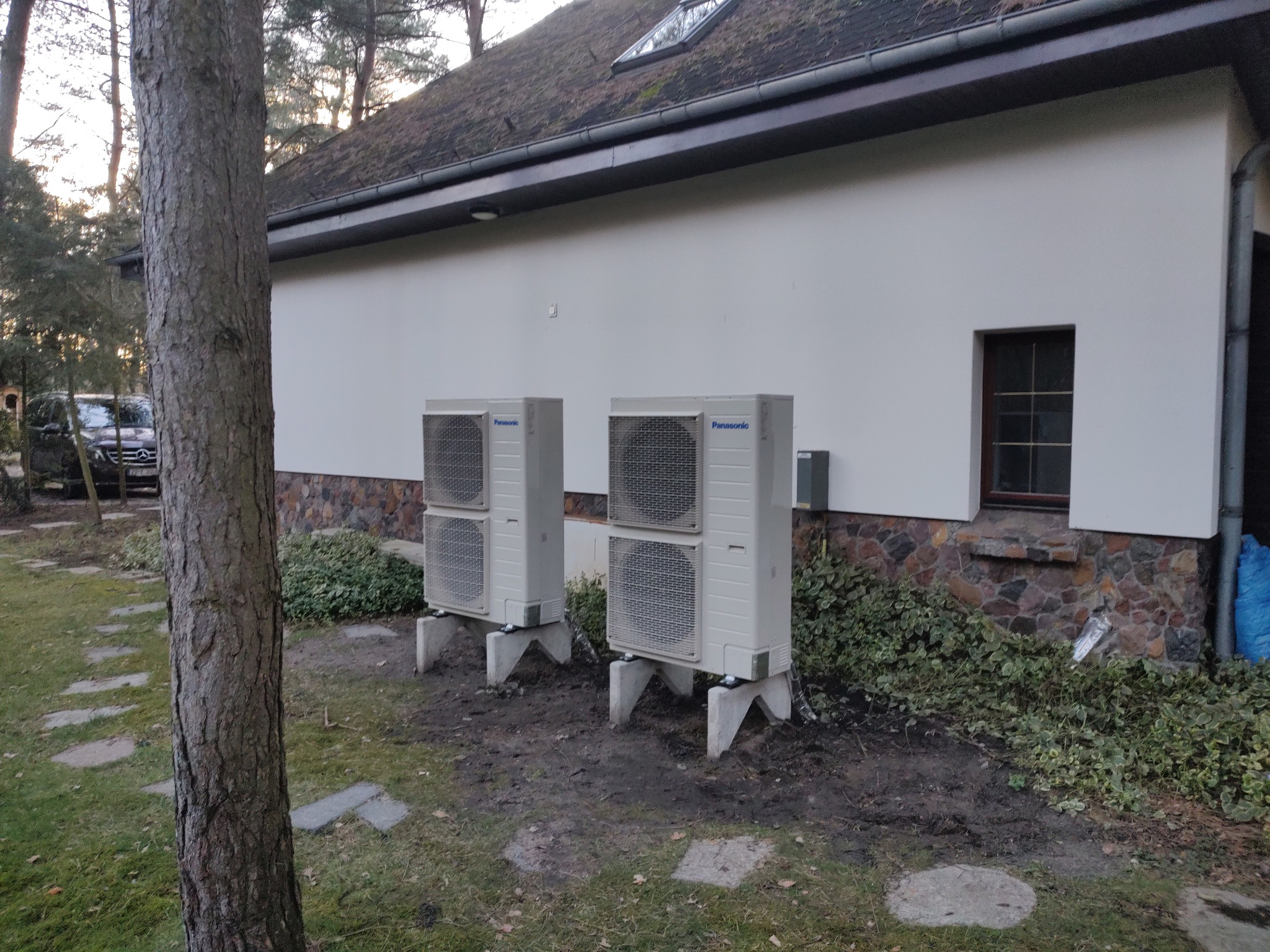 Dwa zewnętrzne moduły pompy ciepła Panasonic ustawione na betonowych podstawach przed budynkiem z białymi ścianami i kamienną podmurówką, widoczne drzewo na pierwszym planie i samochód w tle.