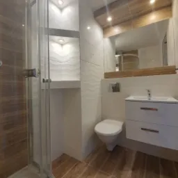 Nowoczesna łazienka z kabiną prysznicową, umywalką z szafką i toaletą; dominują płytki imitujące drewno i białe płytki ze strukturą fal