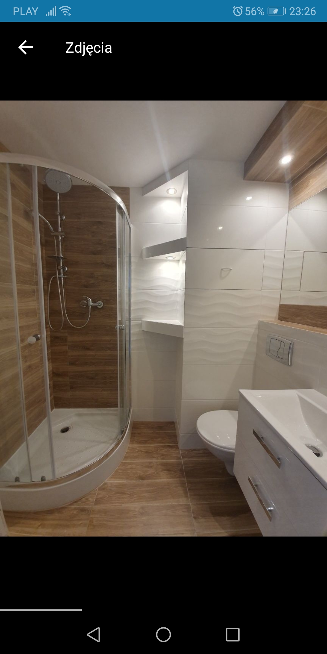 Nowoczesna łazienka z kabiną prysznicową typu walk-in, wykończona płytkami imitującymi drewno na podłodze i wewnątrz kabiny, oraz białymi, falistymi płytkami na ścianach, z podświetlanymi półkami...