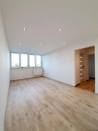 Pusty pokój z panelami podłogowymi w odcieniach beżu, białymi ścianami, oknami z grzejnikiem i wbudowanym oświetleniem punktowym w suficie, widok na otwarte drzwi do innego pomieszczenia.