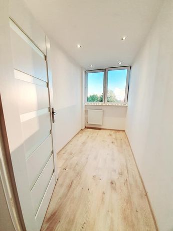 Wąskie, jasne pomieszczenie z drewnopodobną podłogą, białymi ścianami, oknem z widokiem na zieleń i otwartymi białymi drzwiami z poziomymi przeszkleniami.