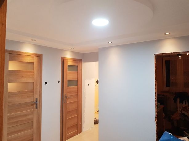 Jasne wnętrze po odświeżeniu, widoczne dwie drewniane drzwi z przeszkleniami, punktowe oświetlenie sufitowe oraz otwór w ścianie przygotowany do montażu ościeżnicy.