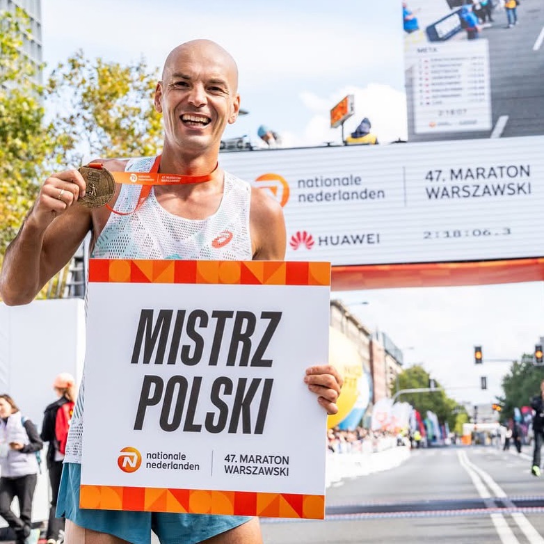 Uśmiechnięty maratończyk z medalem, trzymający tablicę 'Mistrz Polski' na tle mety 47. Maratonu Warszawskiego. Widoczny czas: 2:18:06.3 i logo sponsorów.